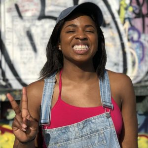 Lanee' Sanders in overalls and a baseball cap smiling big while throwing up a peace sign