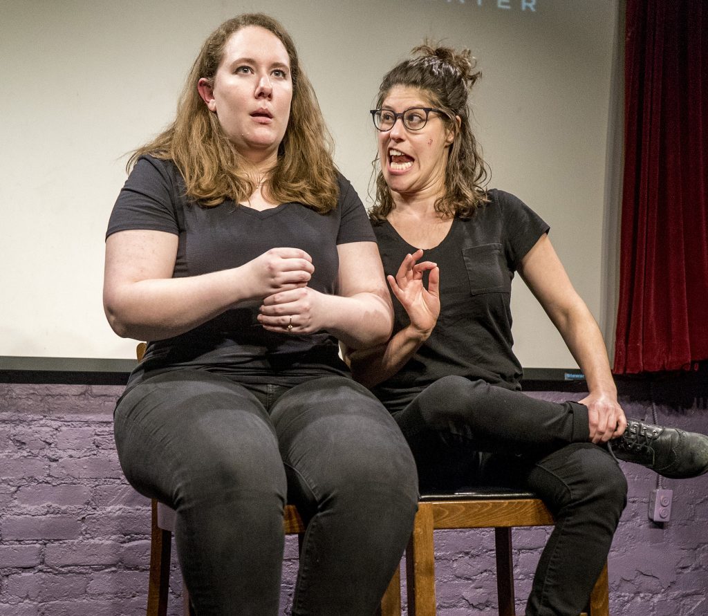 Elena Skopetos and Elana Fishbein seated onstage performing a scene.